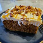 A close-up of a slice of Pumpkin Caramel Pecan Tres Leches Cake, showing its moist texture, creamy whipped topping, and a drizzle of caramel sauce with chopped pecans.