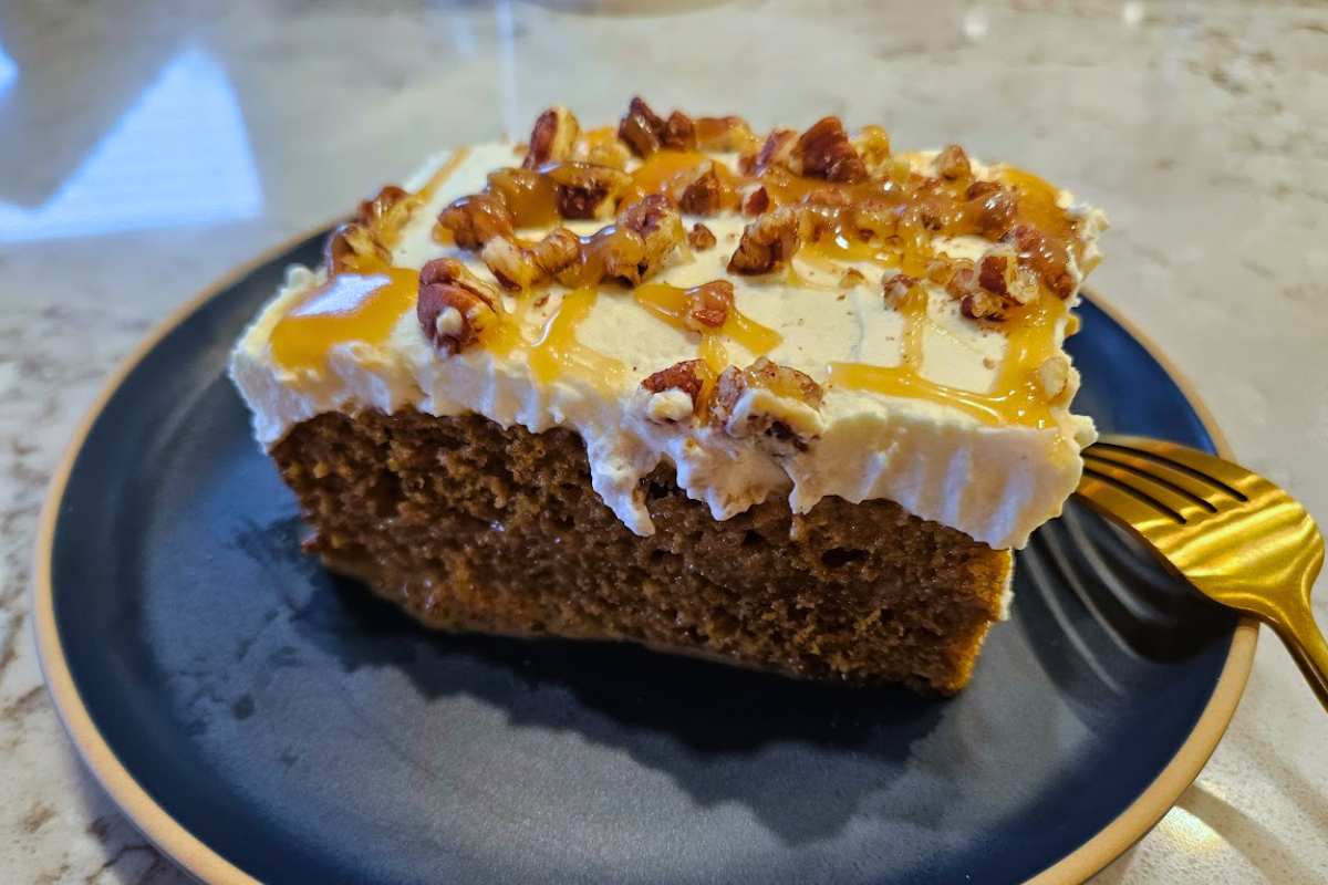 A close-up of a slice of Pumpkin Caramel Pecan Tres Leches Cake, showing its moist texture, creamy whipped topping, and a drizzle of caramel sauce with chopped pecans.